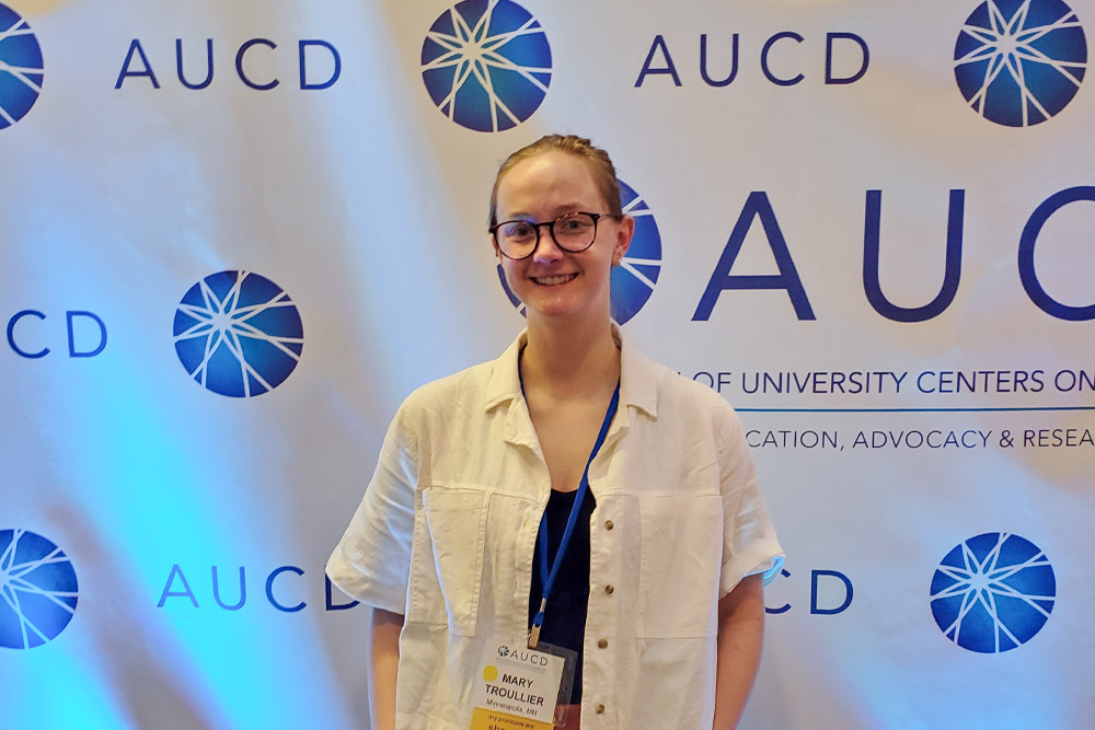 A person stands in front of an AUC D event backdrop with repeating blue logos, wearing glasses, a white shirt over a dark top, and a lanyard badge.