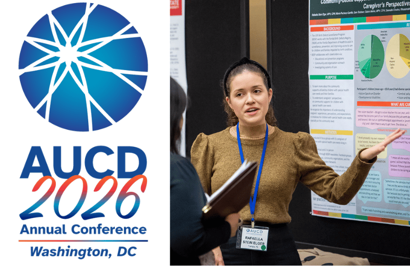 Conference banner reading “AUCD 2026 Annual Conference, Washington, DC” beside a presenter at a booth, gesturing with one hand and holding a folder while standing in front of a large display board.