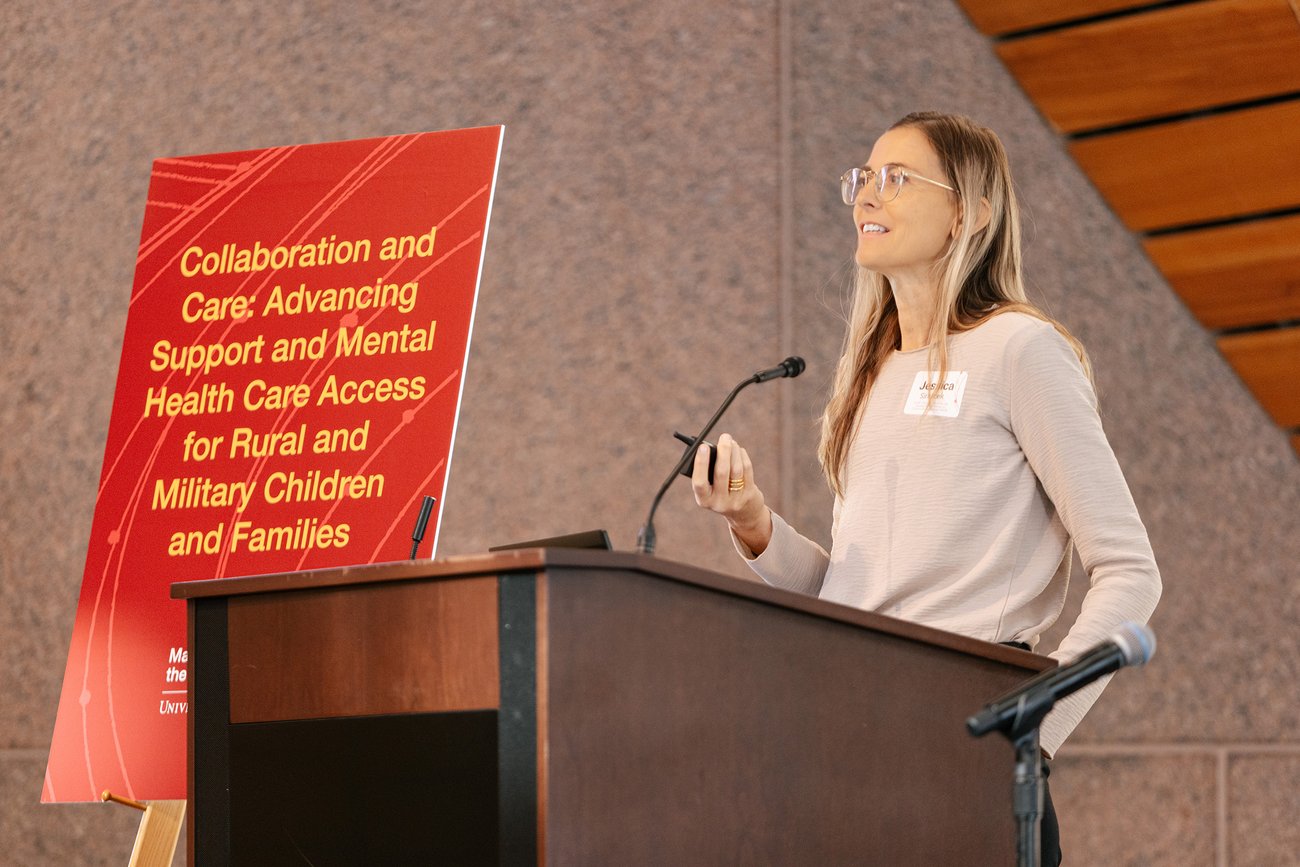 A speaker in a white top addresses an audience at a podium with a microphone, with a red banner behind her reading “Collaboration and Care: Advancing Support and Mental Health Care Access for Rural and Military Children and Families.”